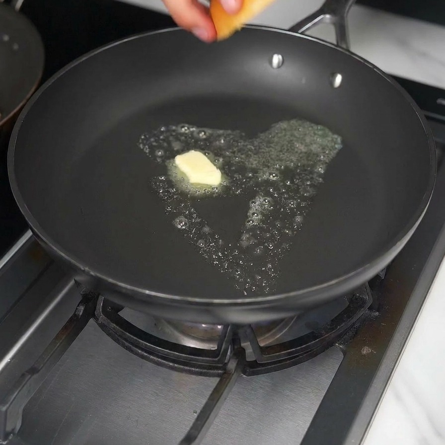 Una mano colocando un pan de hamburguesa tipo brioche en una sartén con un cubo de mantequilla amarilla derritiéndose.