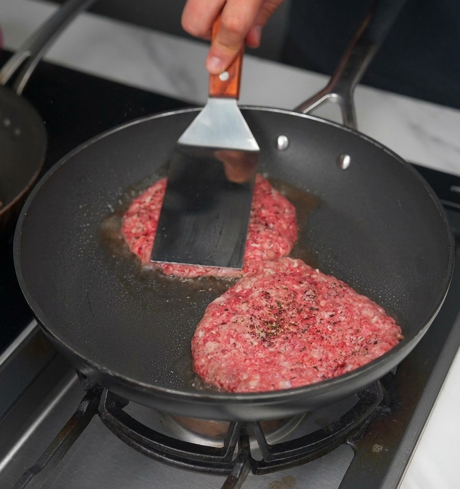 Una espátula metálica presionando dos medallones de carne cruda en una sartén negra para crear una costra.