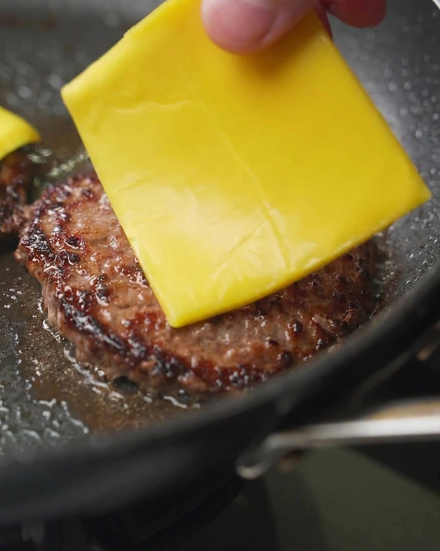 Primer plano de una mano colocando una loncha cuadrada de queso cheddar amarillo sobre un medallón de carne dorado en la sartén.