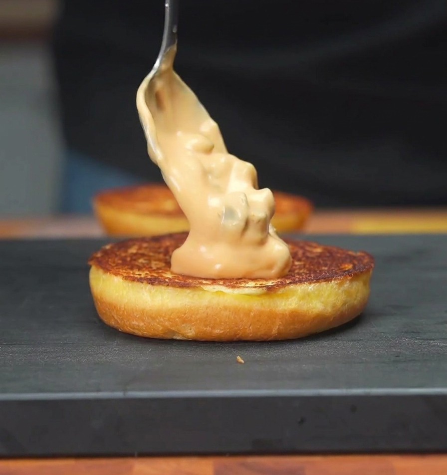 Una cuchara extendiendo una salsa de pepinillos de color naranja cremoso sobre la superficie tostada de un pan de hamburguesa.