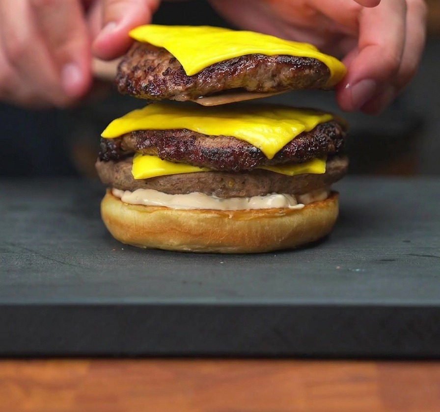Una mano colocando una torre de dos medallones de hamburguesa con queso fundido sobre el pan con salsa.
