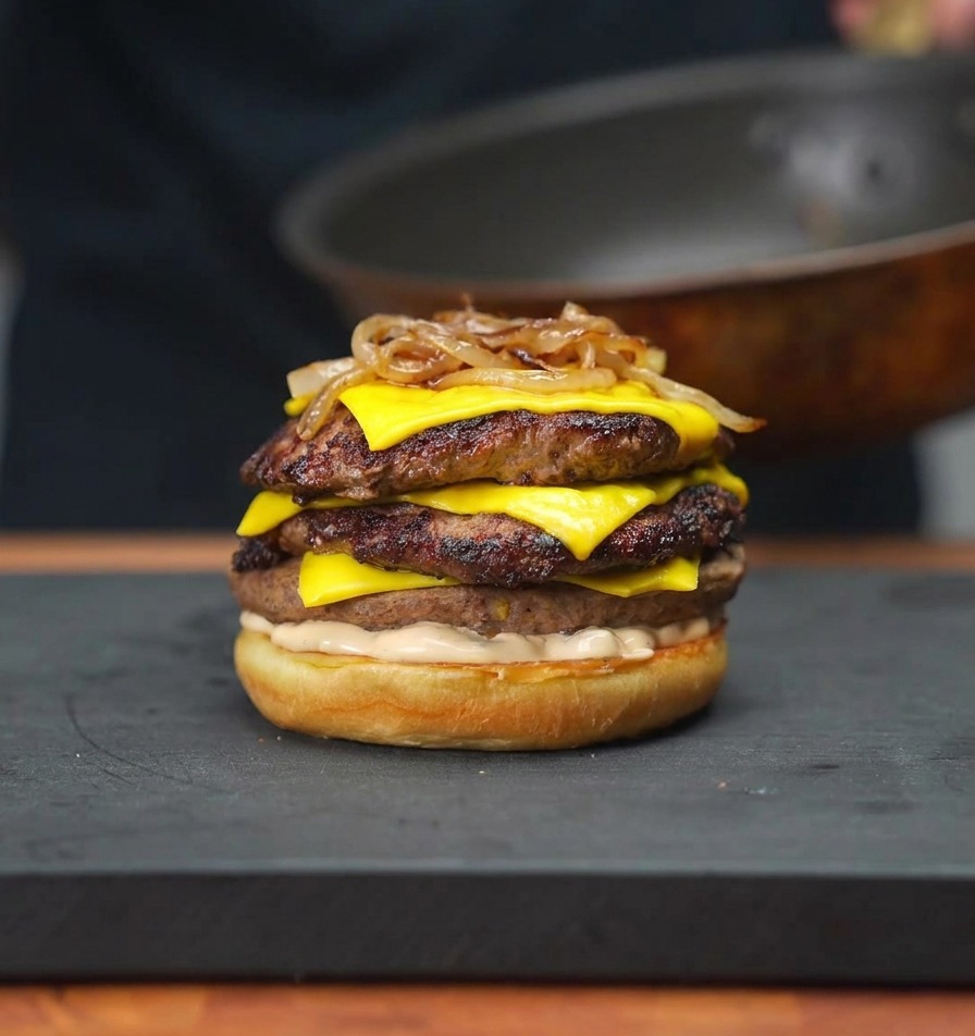 Cebollas caramelizadas siendo colocadas sobre una torre de hamburguesas con queso usando un par de palillos.
