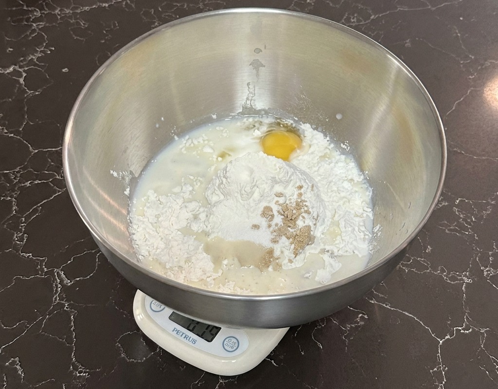 Un bol de acero inoxidable sobre una báscula digital que contiene harina, leche, un huevo y levadura.