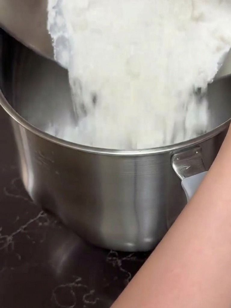Manos vertiendo una mezcla de harina blanca y leche desde un bol de acero a un bol de batidora de pie grande.