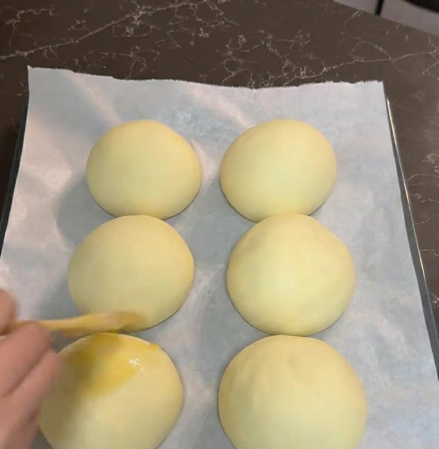 Una mano usando un pincel de pastelería para cubrir seis bolas de masa levadas con huevo batido en una bandeja con papel vegetal.