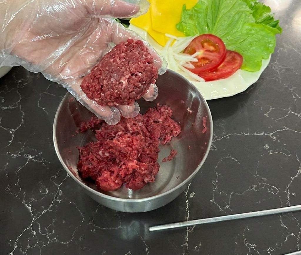 Una mano con guante de plástico sosteniendo una bola de carne picada cruda sobre un bol de acero inoxidable.