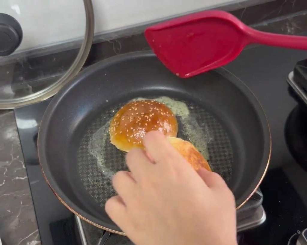 Mitades de pan de hamburguesa tostándose boca abajo en una sartén con mantequilla derretida burbujeante.