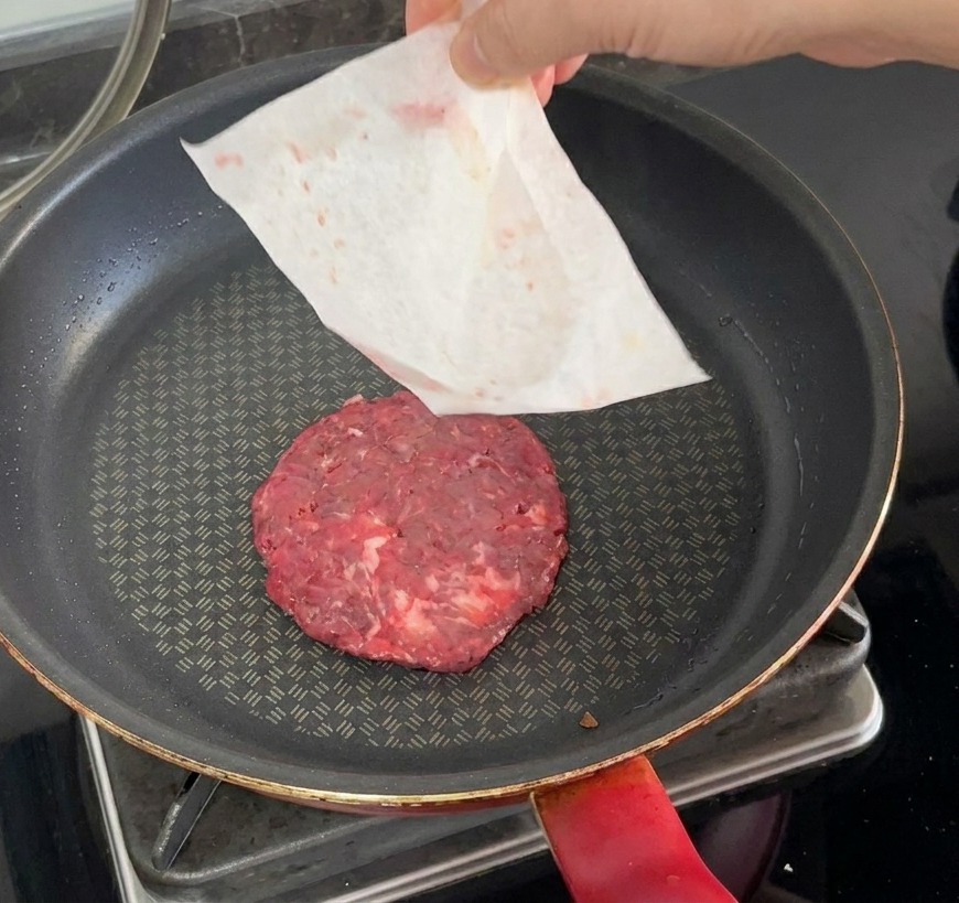 Una hamburguesa cruda siendo colocada en una sartén caliente mientras se retira el papel vegetal con la mano.