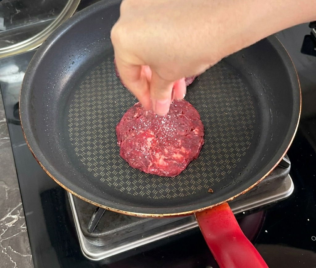 Una mano espolvoreando sal sobre una hamburguesa que se está sellando en una sartén antiadherente oscura.