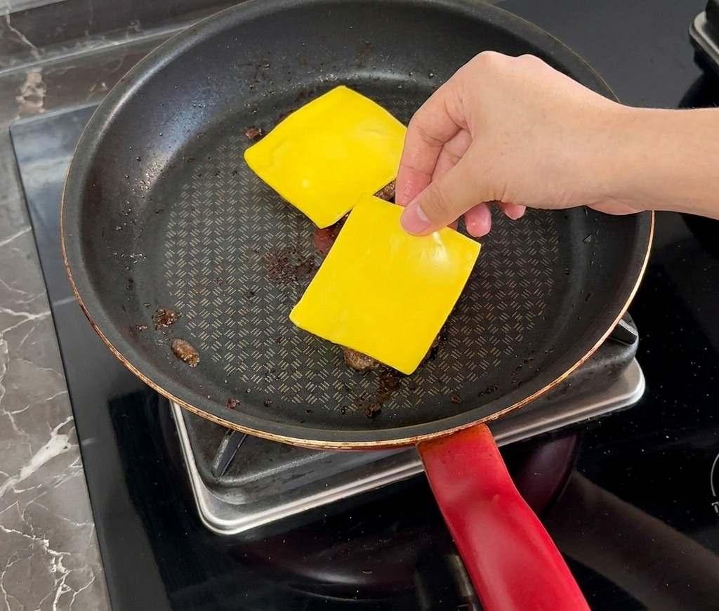 Una mano colocando una loncha cuadrada de queso amarillo sobre una hamburguesa cocinada en la sartén.