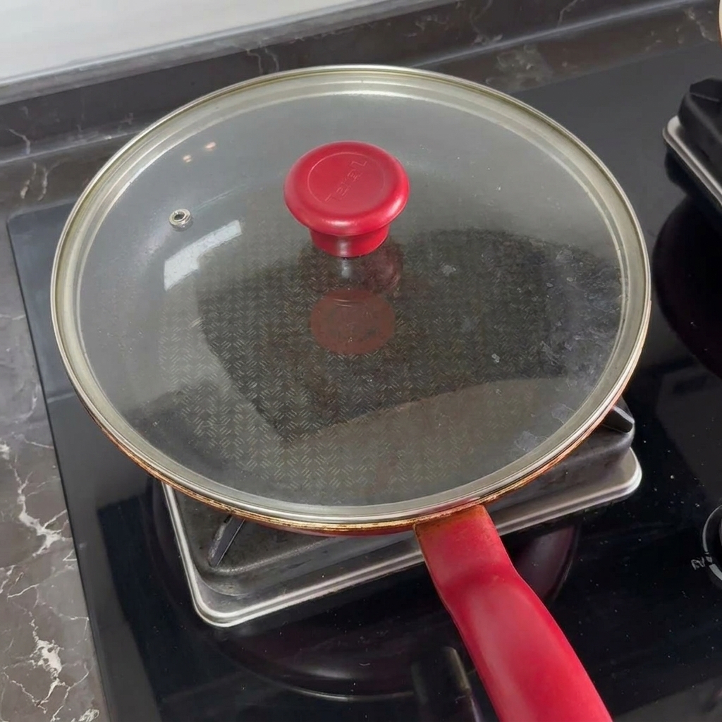 Una tapa de vidrio con mango rojo cubriendo una sartén para fundir el queso sobre las hamburguesas.