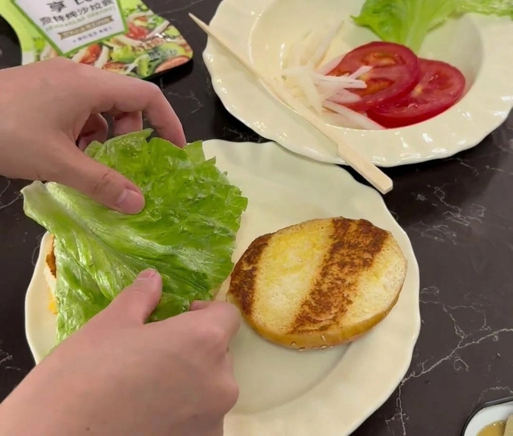 Manos colocando una hoja de lechuga verde fresca sobre un pan de hamburguesa tostado en un plato blanco.