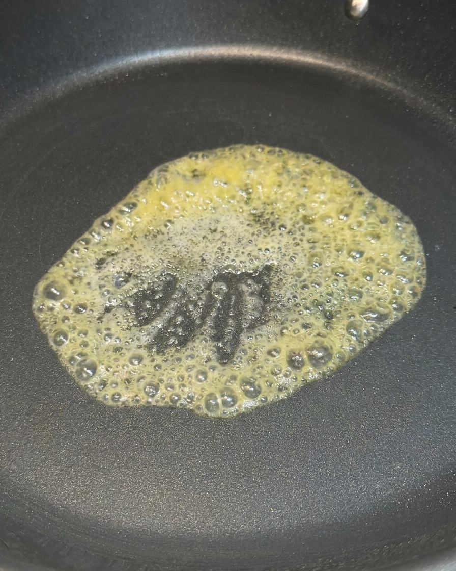 Un trozo de mantequilla amarilla derritiéndose y burbujeando en el centro de una sartén antiadherente negra.