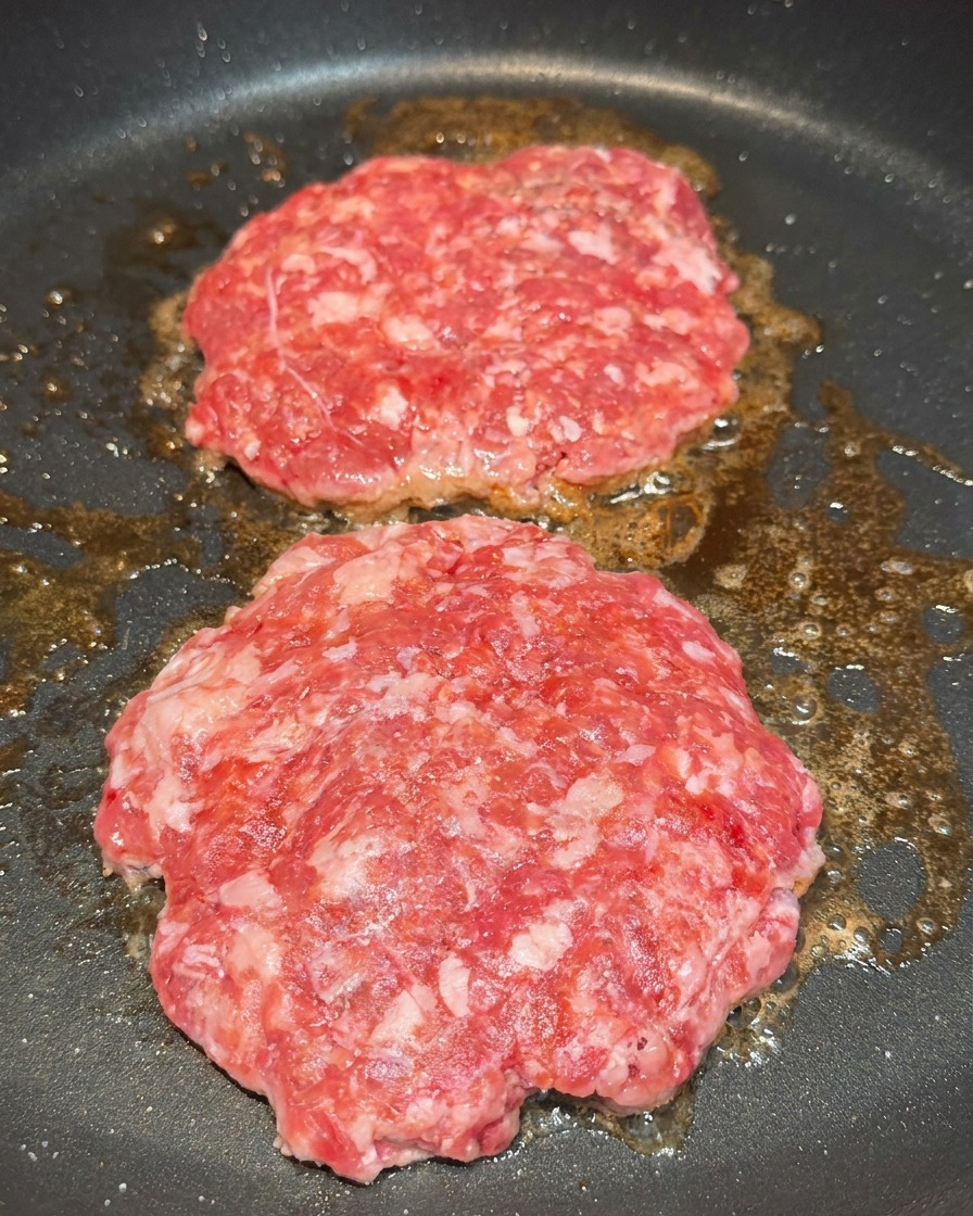 Dos medallones de carne en la sartén siendo sazonados con granos visibles de sal y pimienta negra.