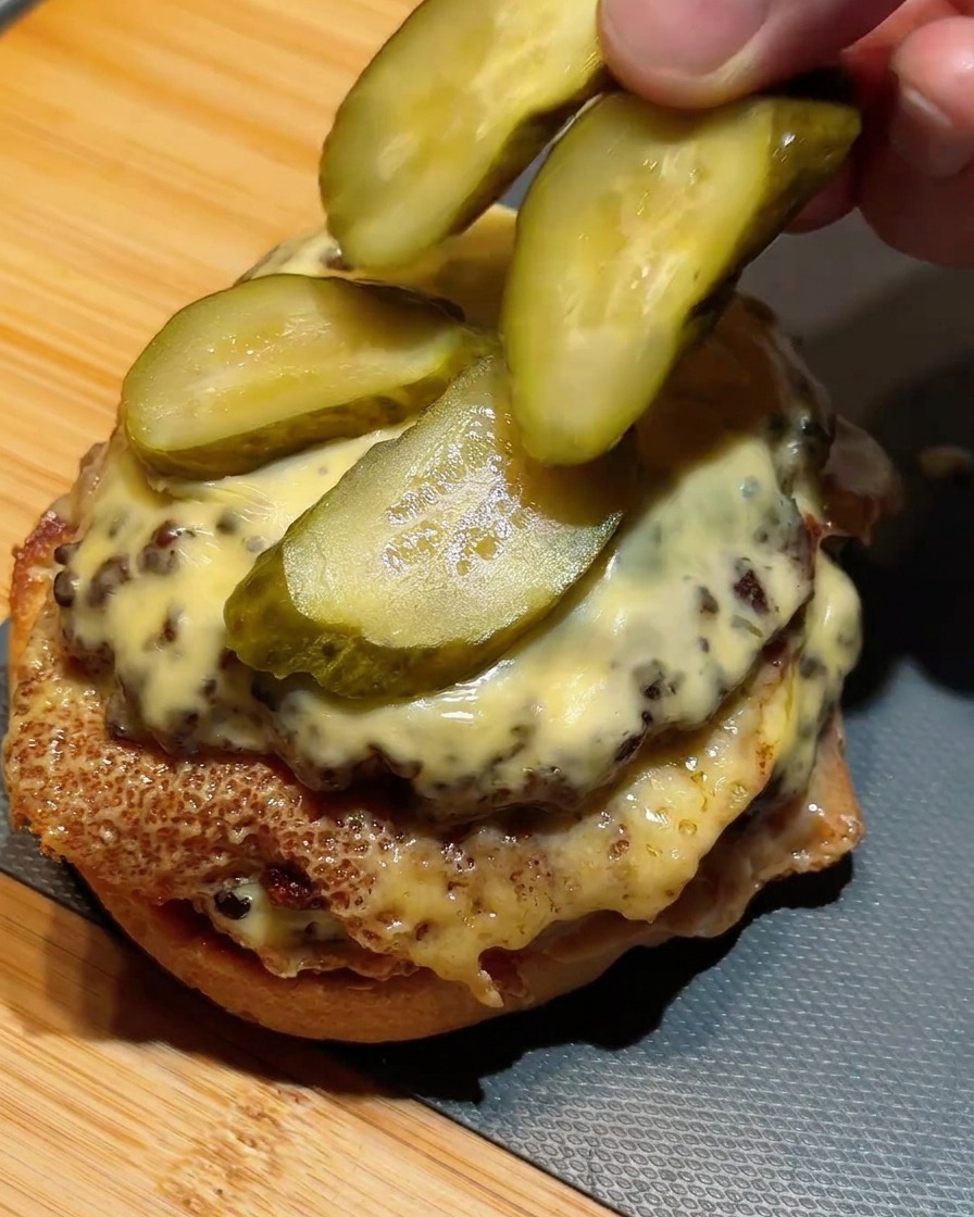 Dedos colocando rodajas de pepinillos verdes sobre una hamburguesa doble con queso fundido.