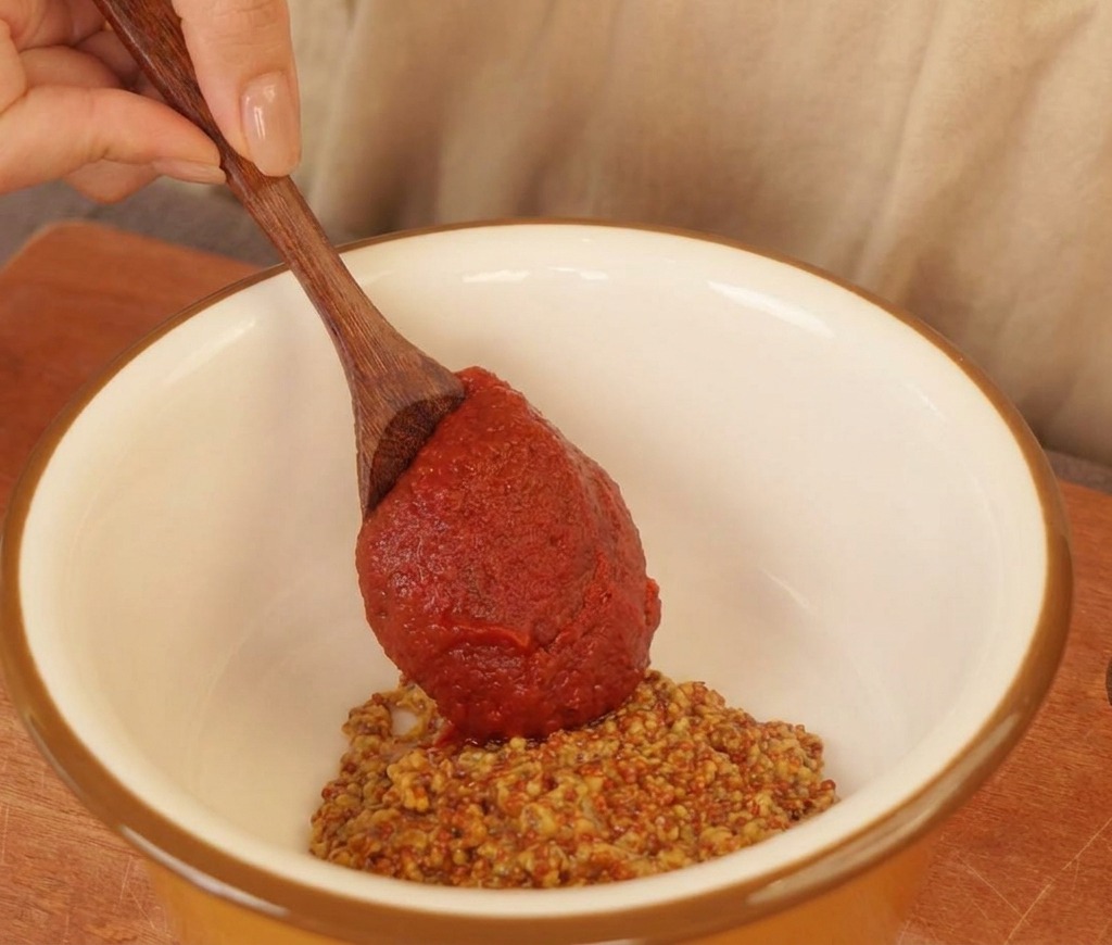 Una cuchara de madera añadiendo salsa de chile roja a un bol blanco con mostaza a la antigua y mayonesa.