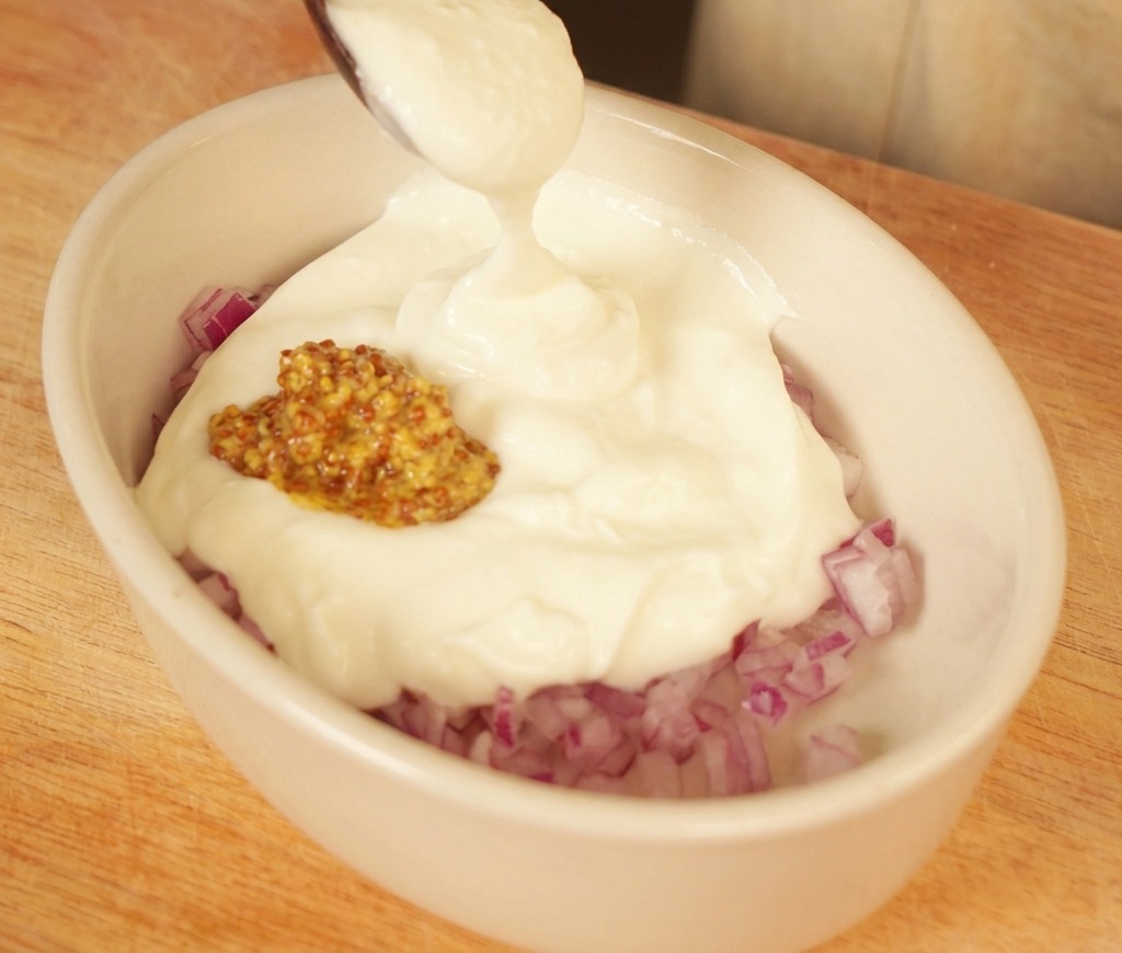 Una cucharada de yogur blanco cremoso añadiéndose a un plato ovalado con cebolla roja picada y mostaza.