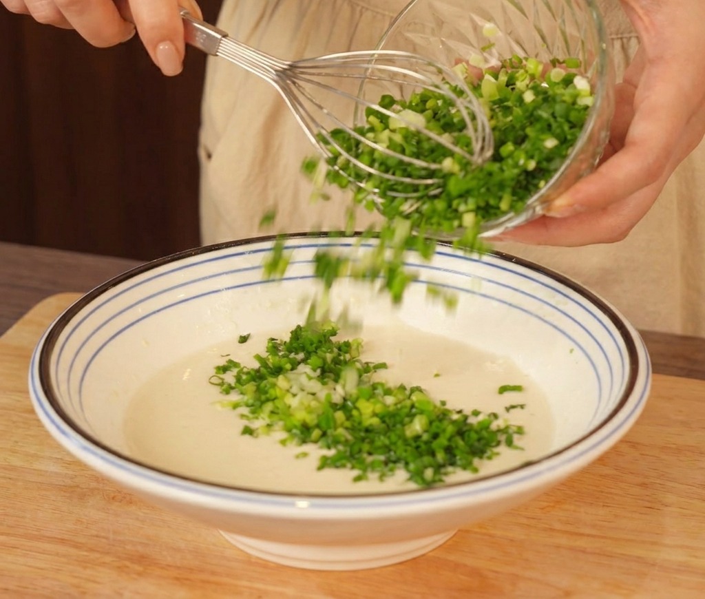 Un bol grande con una masa clara siendo batida mientras se añade una cantidad generosa de cebollino verde picado.