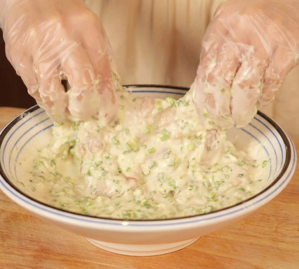 Manos con guantes mezclando muslos de pollo en un bol de masa blanca espesa llena de cebollino picado.