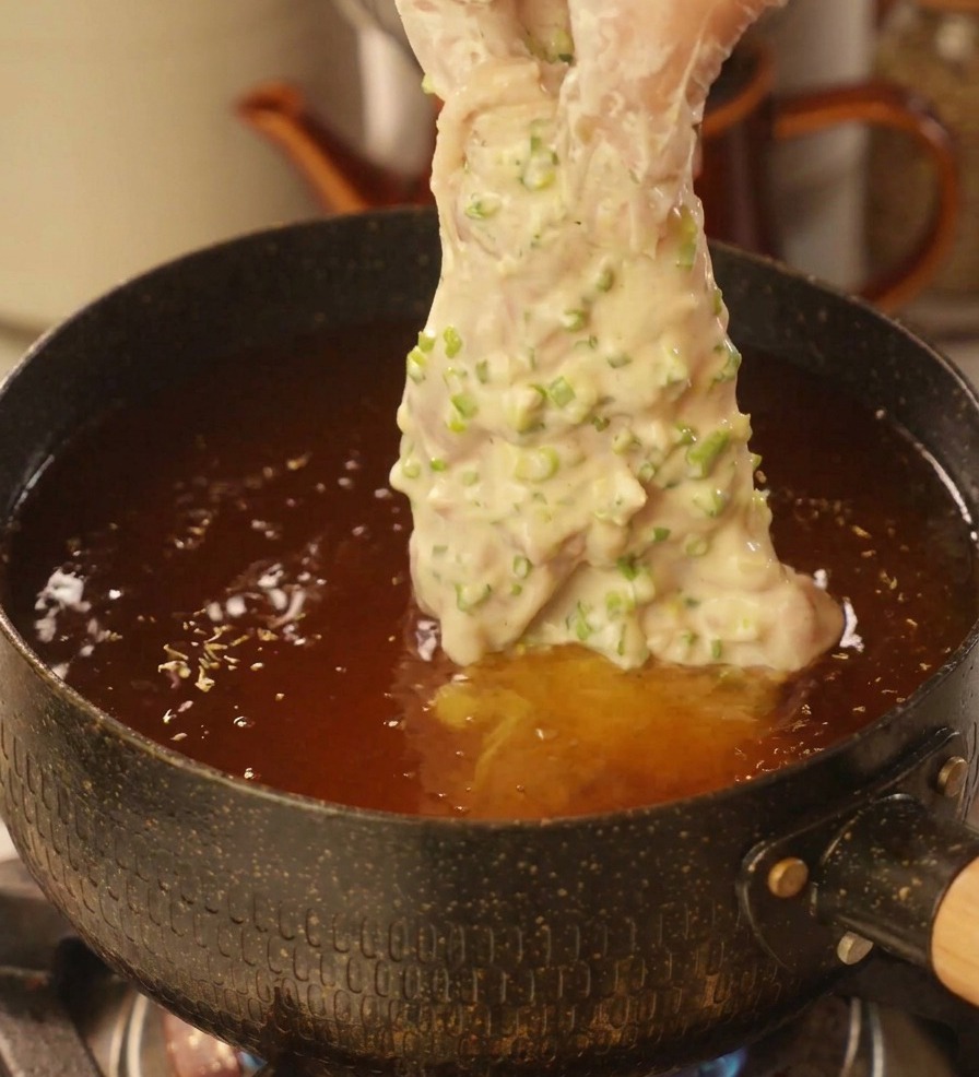Una mano con guante de plástico introduciendo un trozo de pollo cubierto de masa de cebollino en una olla con aceite caliente.