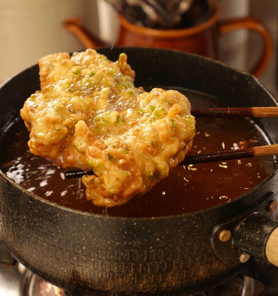 Pollo frito grande y dorado saliendo de la olla con aceite mediante unos palillos de madera oscuros.