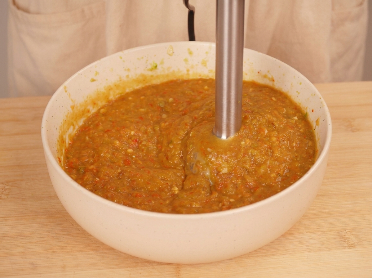 Una batidora de mano triturando verduras asadas dentro de un bol blanco para crear una salsa espesa y texturizada de color naranja verdoso.