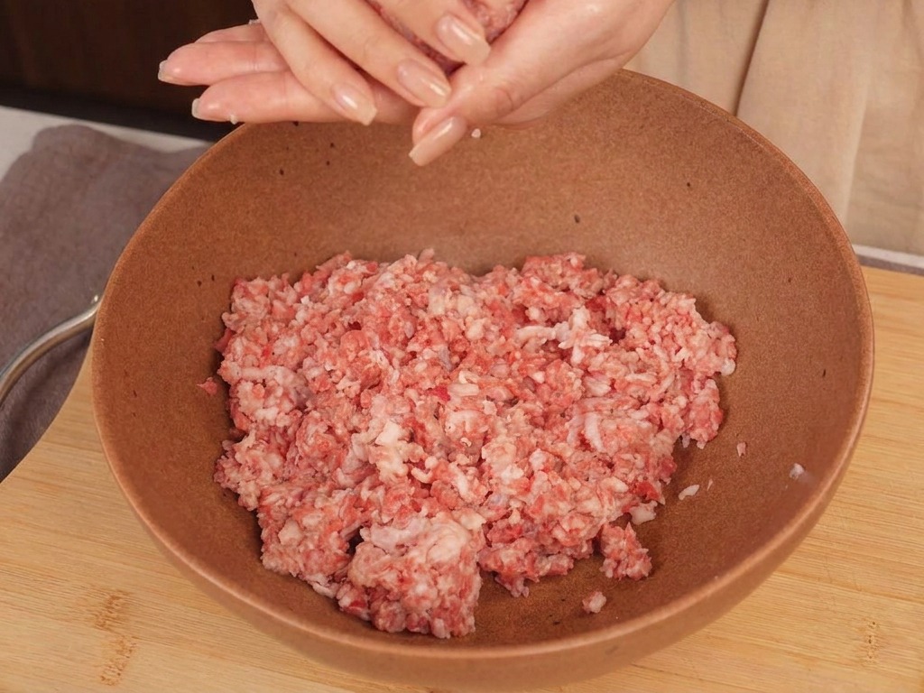 Las manos de una persona dando forma a una bola redonda de carne picada cruda sobre un bol de cerámica.