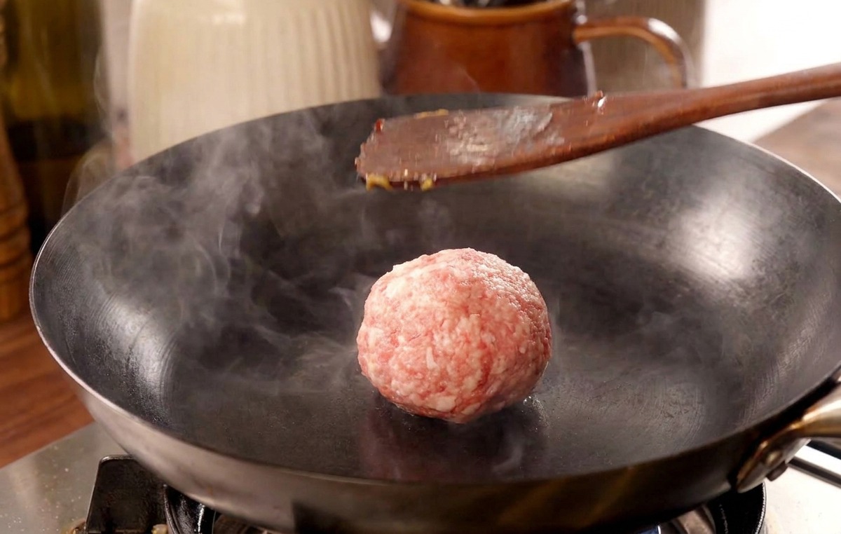 Una espátula de madera presionando una bola de carne en una sartén muy caliente, liberando una nube de vapor.