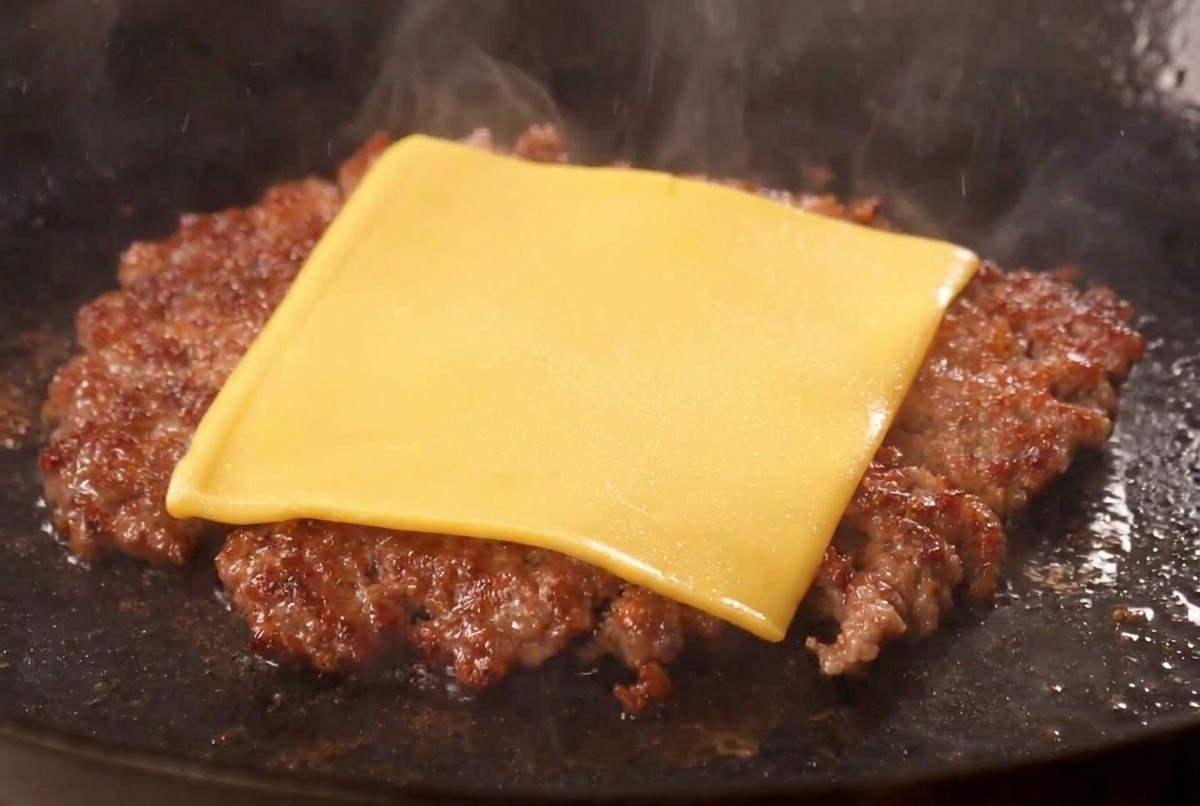 Una loncha de queso amarillo americano fundiéndose sobre la parte superior de una hamburguesa sellada con costra en la sartén.