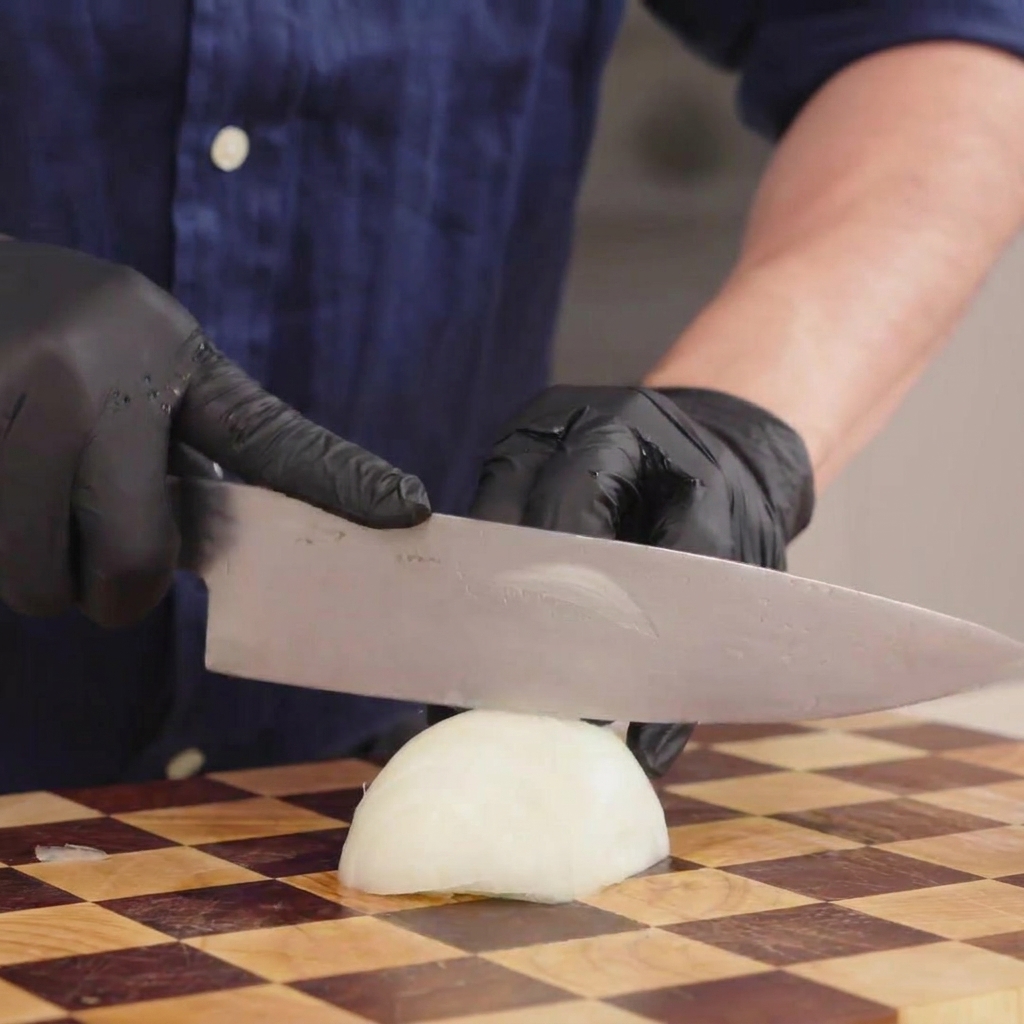 Primer plano de una persona con guantes negros cortando una cebolla blanca en rodajas finas sobre una tabla de madera.