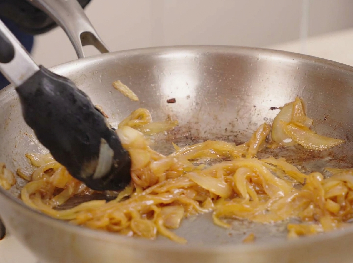 Pinzas removiendo una mezcla espesa y dorada de cebollas caramelizadas en una sartén brillante.