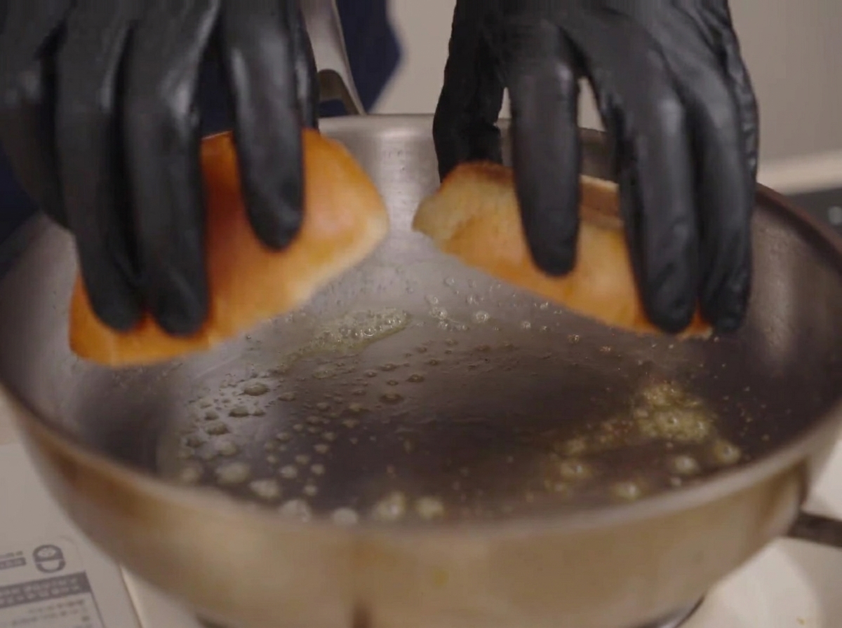 Manos con guantes colocando dos mitades de pan brioche en una sartén con mantequilla derretida burbujeante.