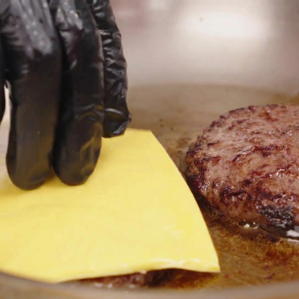 Mano con guante colocando una loncha de queso amarillo sobre una hamburguesa de carne caliente en la sartén.