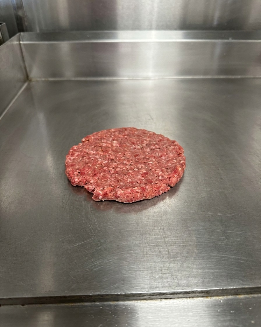 Una hamburguesa aplastada en la plancha, sazonada con granos visibles de sal y pimienta.