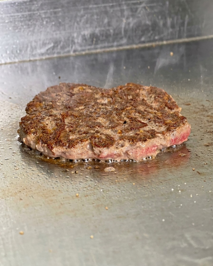 Una hamburguesa volteada en la plancha mostrando una costra marrón oscuro y crujiente con grasa burbujeando.