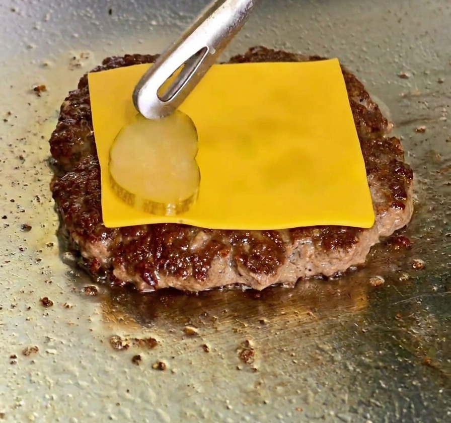 Una rebanada de queso cheddar fundido sobre la carne mientras unas pinzas colocan una rodaja de pepinillo encima.