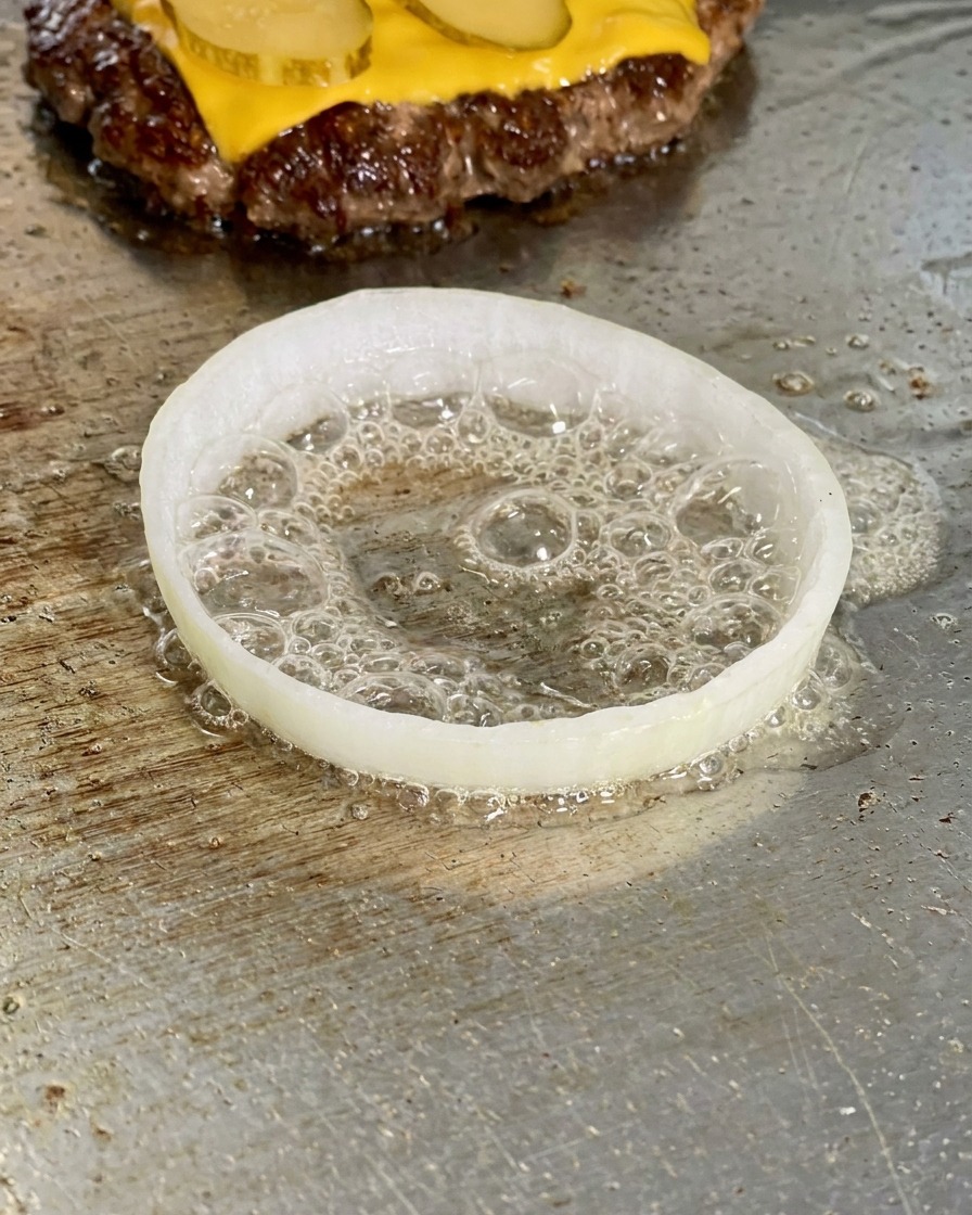 Una rodaja de cebolla blanca en la plancha con un huevo crudo friéndose dentro, rodeado de aceite burbujeante.