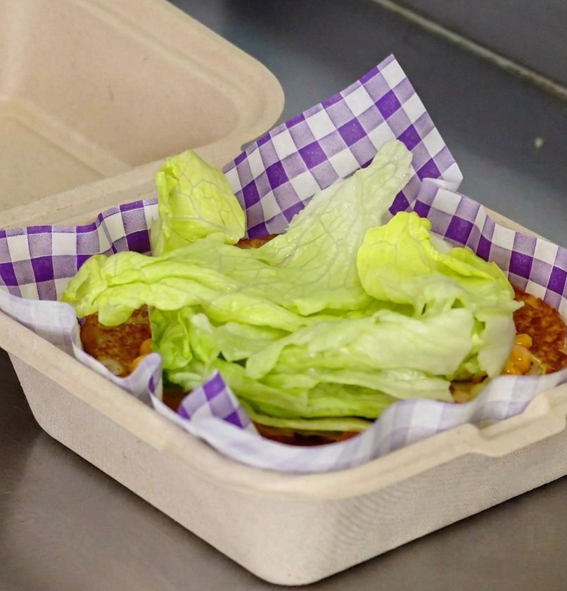 Hojas frescas de lechuga verde apilándose sobre un pan tostado con salsa roja dentro de un papel cuadriculado.