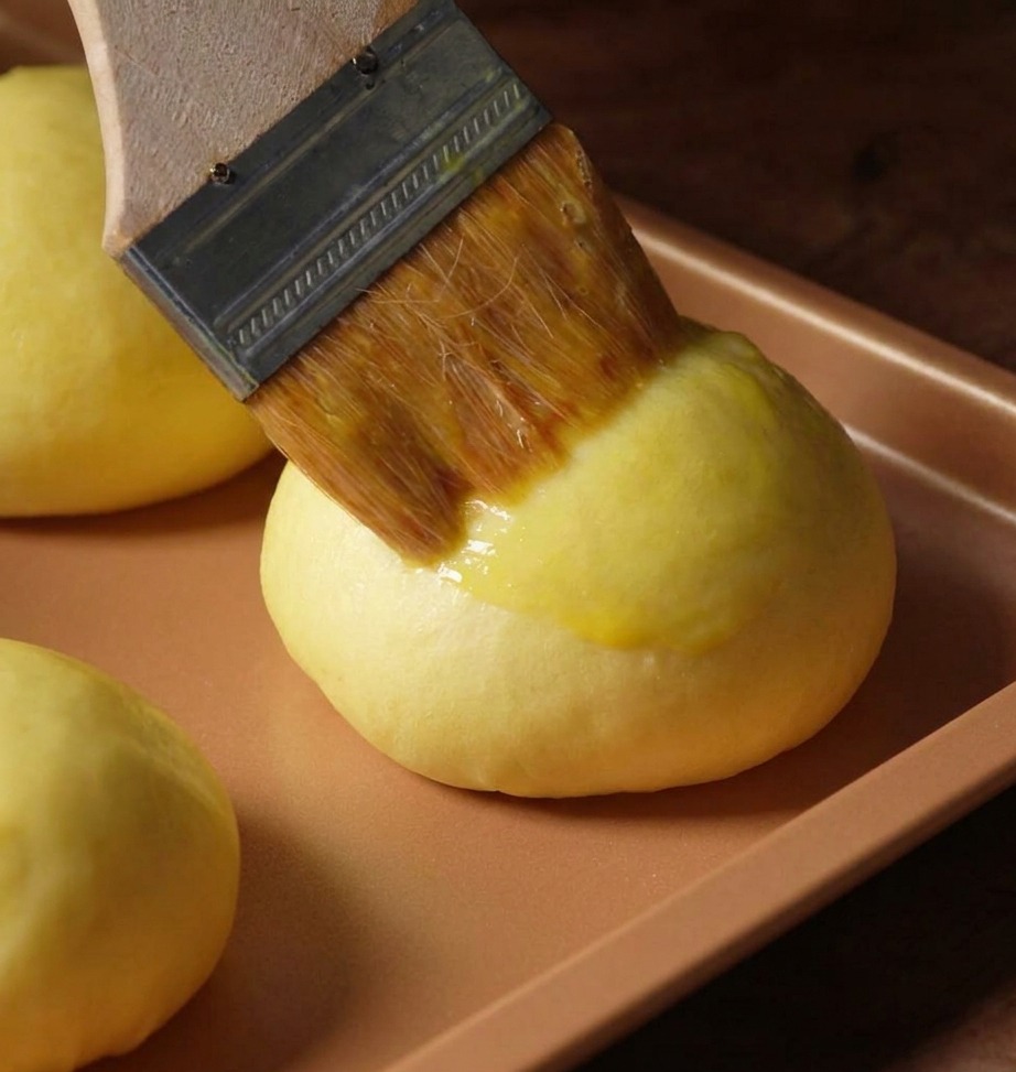 Pincel de pastelería aplicando barniz de huevo sobre un bollo de masa en una bandeja de color cobre.