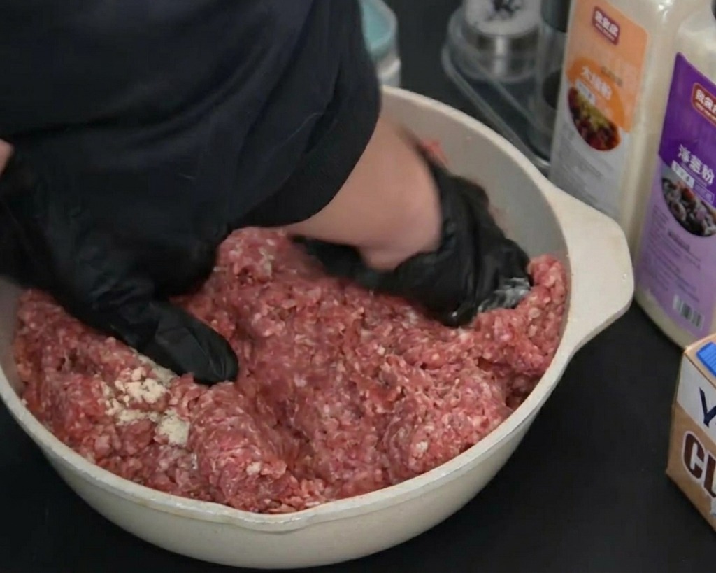 Manos con guantes negros mezclando y amasando la carne molida sazonada en una bandeja clara.