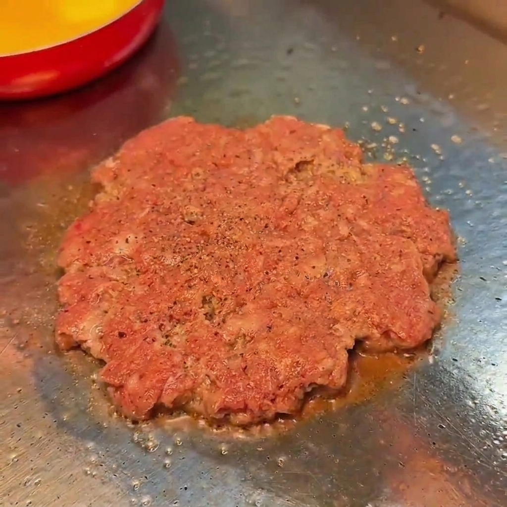 Un disco de carne de ternera cocinándose en una plancha, sazonado con sal y pimienta negra.