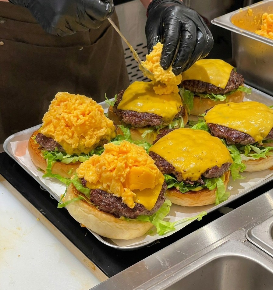 Un chef usando una cuchara para colocar una gran porción de huevos revueltos sobre las hamburguesas.