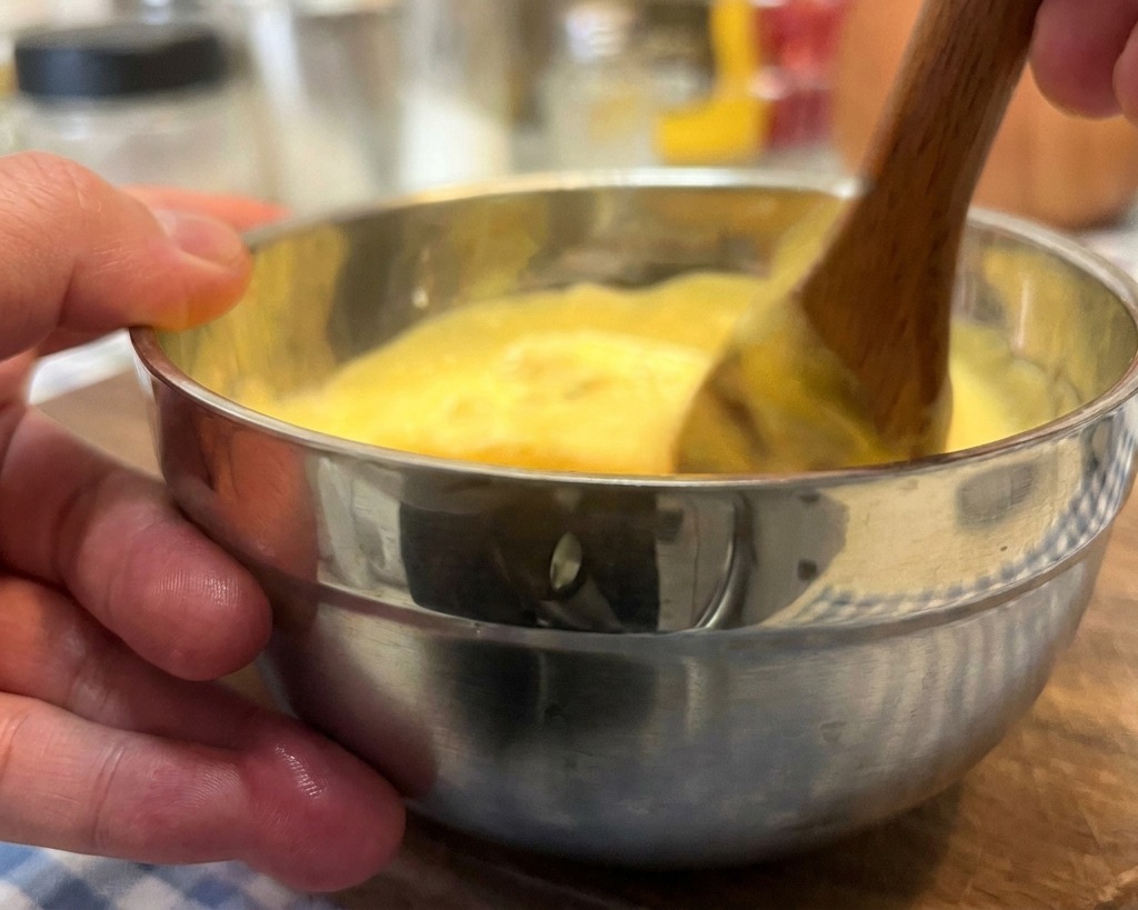 Mano removiendo una salsa cremosa de tono amarillento con una cuchara de madera en un bol de acero.
