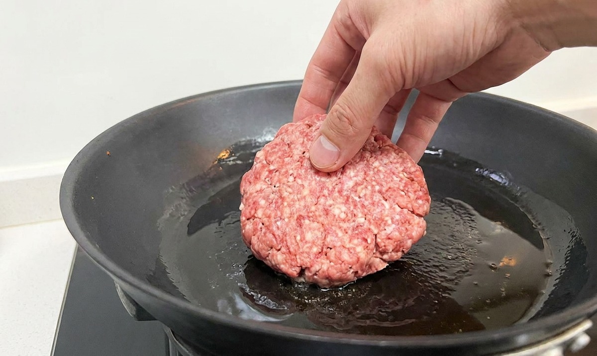 Mano colocando un disco de carne picada en una sartén de hierro caliente con aceite brillante.