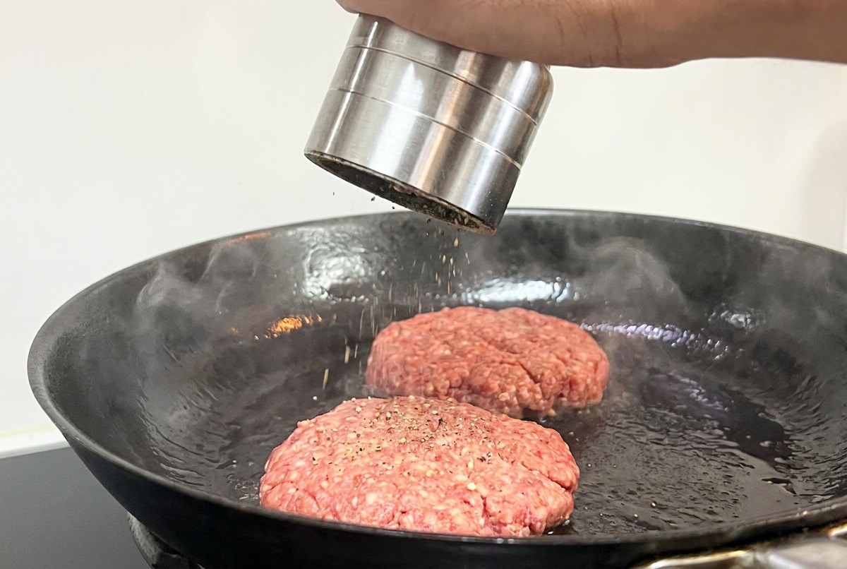 Mano usando un molinillo de pimienta para sazonar dos hamburguesas que se cocinan en la sartén.