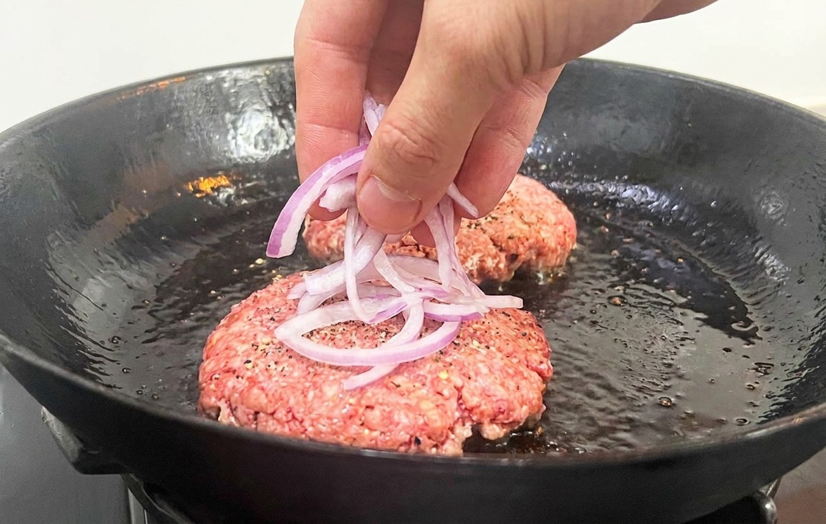 Mano poniendo finas tiras de cebolla sobre la carne cruda en la sartén.