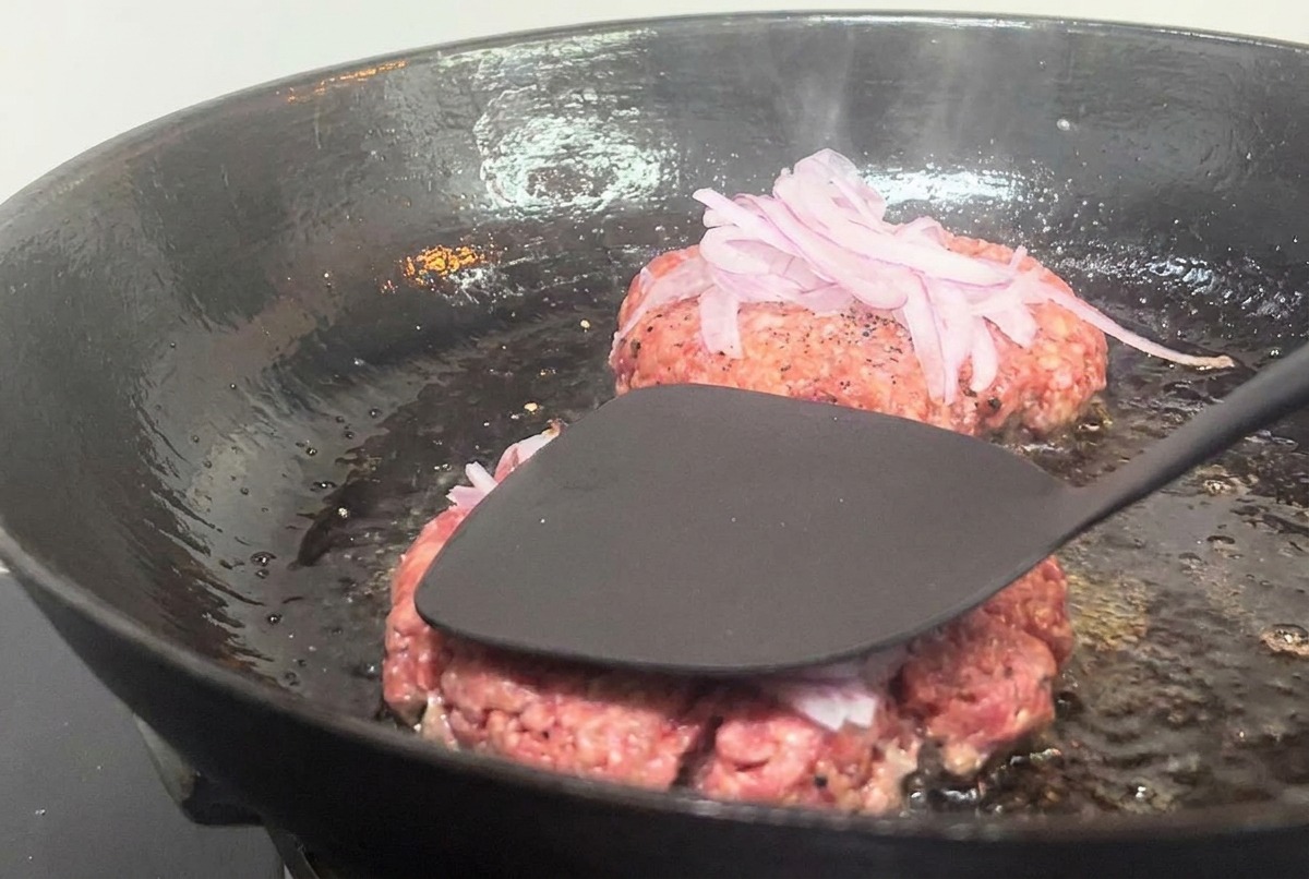 Espátula metálica presionando con fuerza una hamburguesa con cebolla en una sartén caliente.