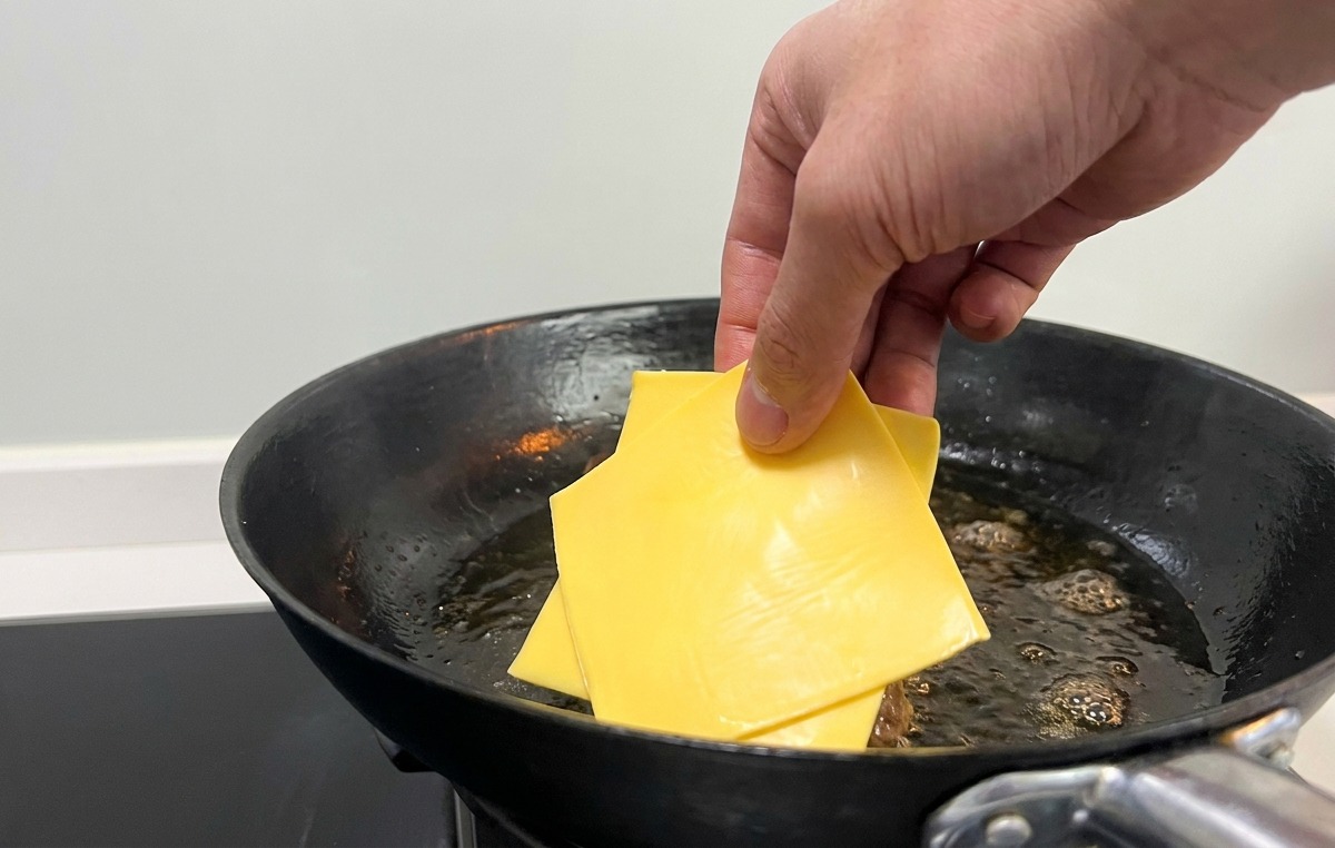 Mano colocando lonchas de queso amarillo sobre la carne sellada en la sartén.