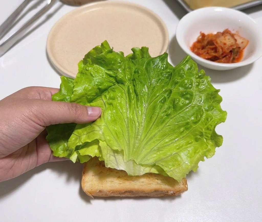 Una mano colocando una hoja de lechuga verde sobre una tostada en una encimera blanca.