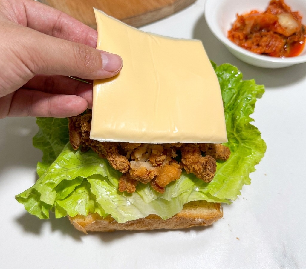 Una mano colocando una loncha de queso sobre el pollo frito y la lechuga.